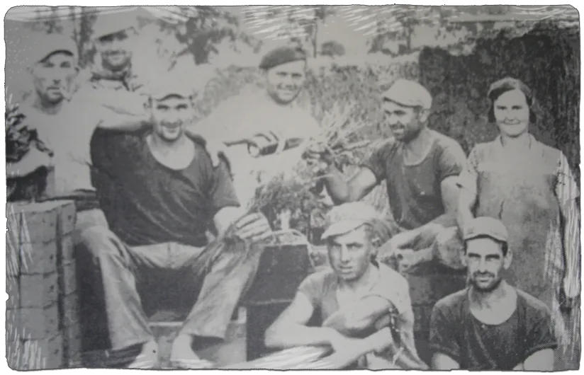 Vintage zwart-witfoto van acht werkers in platte petten en schorten, poserend bij kisten met geoogste groenten